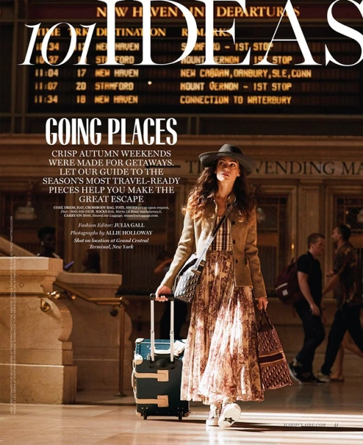 Editorial fashion shot of woman with luggage in a train station with arrival and departure boards, natural lighting, stylish travel attire.