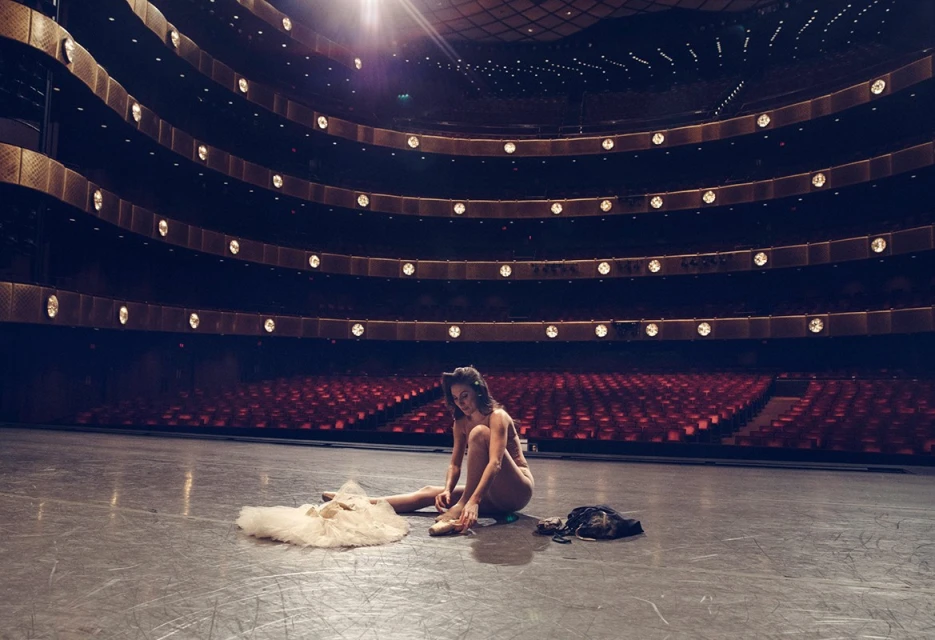 Ballerina sits on a stage tying her ballet shoes in a theater, with a white tutu and garment bag beside her, with rows of seats.