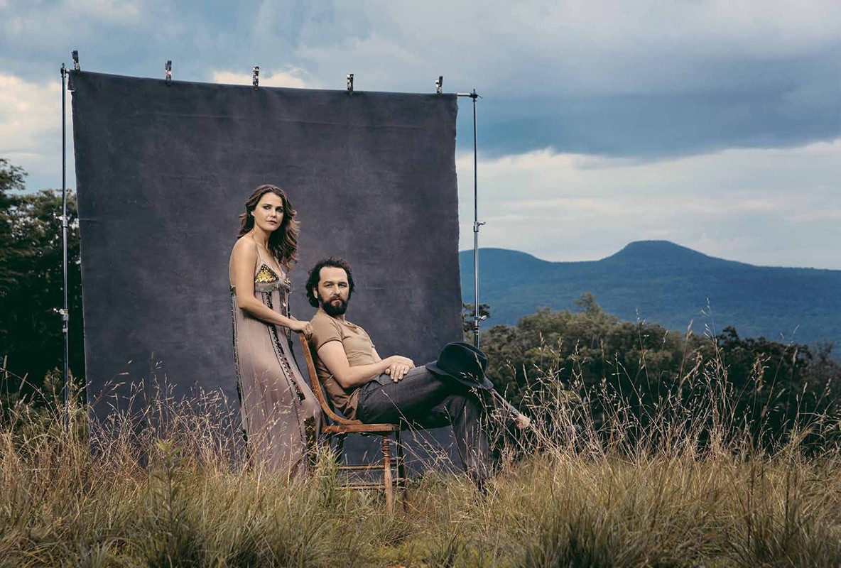 A man seated on a wooden chair and a woman standing beside him outdoors before a dark backdrop with mountains behind them.
