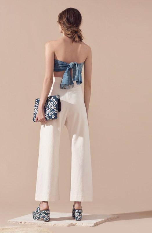Rear view of a model in a blue strapless top, white trousers, and platform shoes holding a patterned clutch.