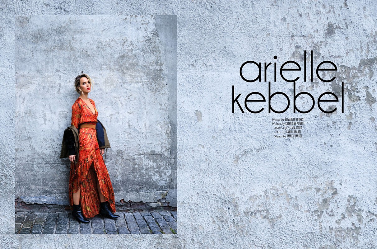 Model Arielle Kebbel poses in an orange dress against a weathered wall backdrop, adding a jacket, complemented by ankle boots.