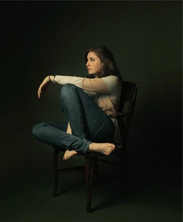 Woman seated barefoot with legs curled on a dark wooden chair against a dark moody green backdrop.