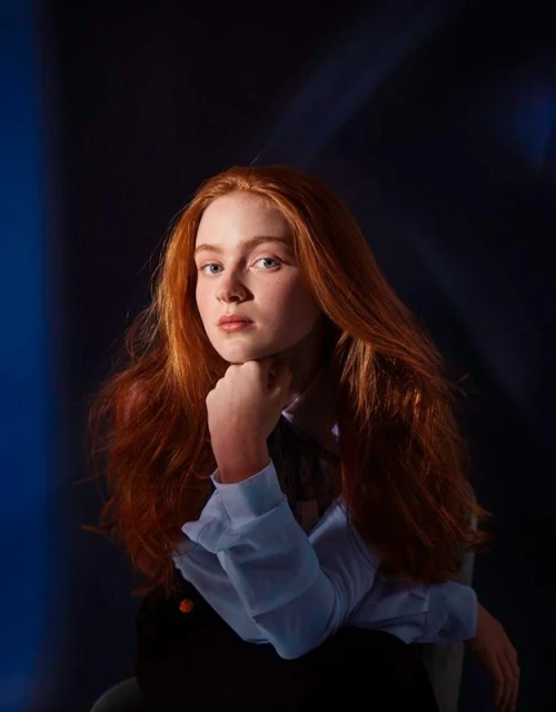 Close-up portrait of a red-haired young woman with a hand near her chin, wearing a light blue blouse and dark pants against a dark background.