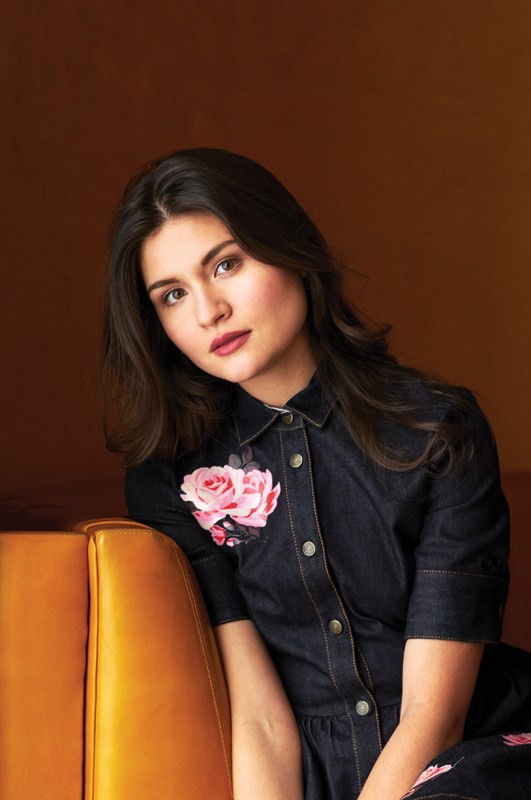 Portrait of a woman with dark hair wearing a dark denim dress with floral accents, leaning against a vintage-style orange couch.