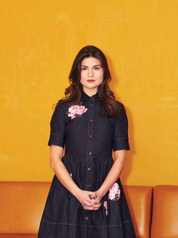 Portrait of a brunette woman in a denim dress with floral accents standing against a yellow wall with a tan couch.