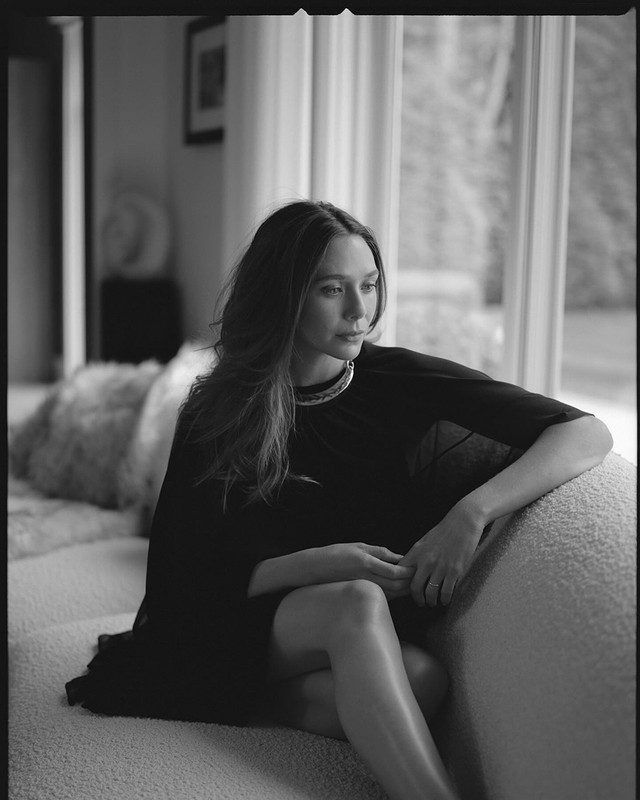 Black and white portrait of a seated woman wearing a dark dress and a silver choker necklace in a naturally lit interior setting.