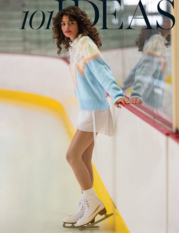 Female figure skater in a pastel sweater poses on the ice rink next to the rink wall, illuminated by bright lighting.
