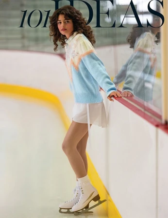 Female figure skater in a pastel sweater poses on the ice rink next to the rink wall, illuminated by bright lighting.