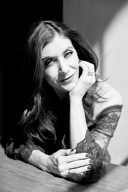 Black and white portrait of a woman with dark hair wearing a lace dress, seated at a light brown wooden table indoors.