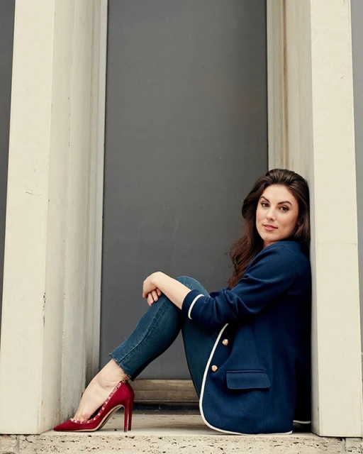 Brunette woman in a blue blazer and jeans sits against a gray wall wearing red high heels; a fashion shot taken outdoors.