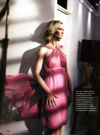 A fashionable adult woman in a formal pink dress and jewelry poses in a bright indoor setting with sunlight and shadows.