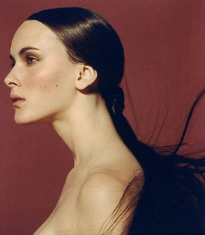 Close-up side portrait of an adult female with long, dark hair in a ponytail against a dark-red background, showcasing beauty and style.