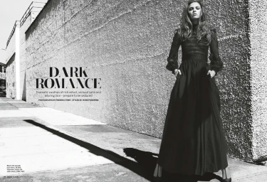 Model standing outdoors wearing a long black gown against a textured concrete wall under bright sunlight.