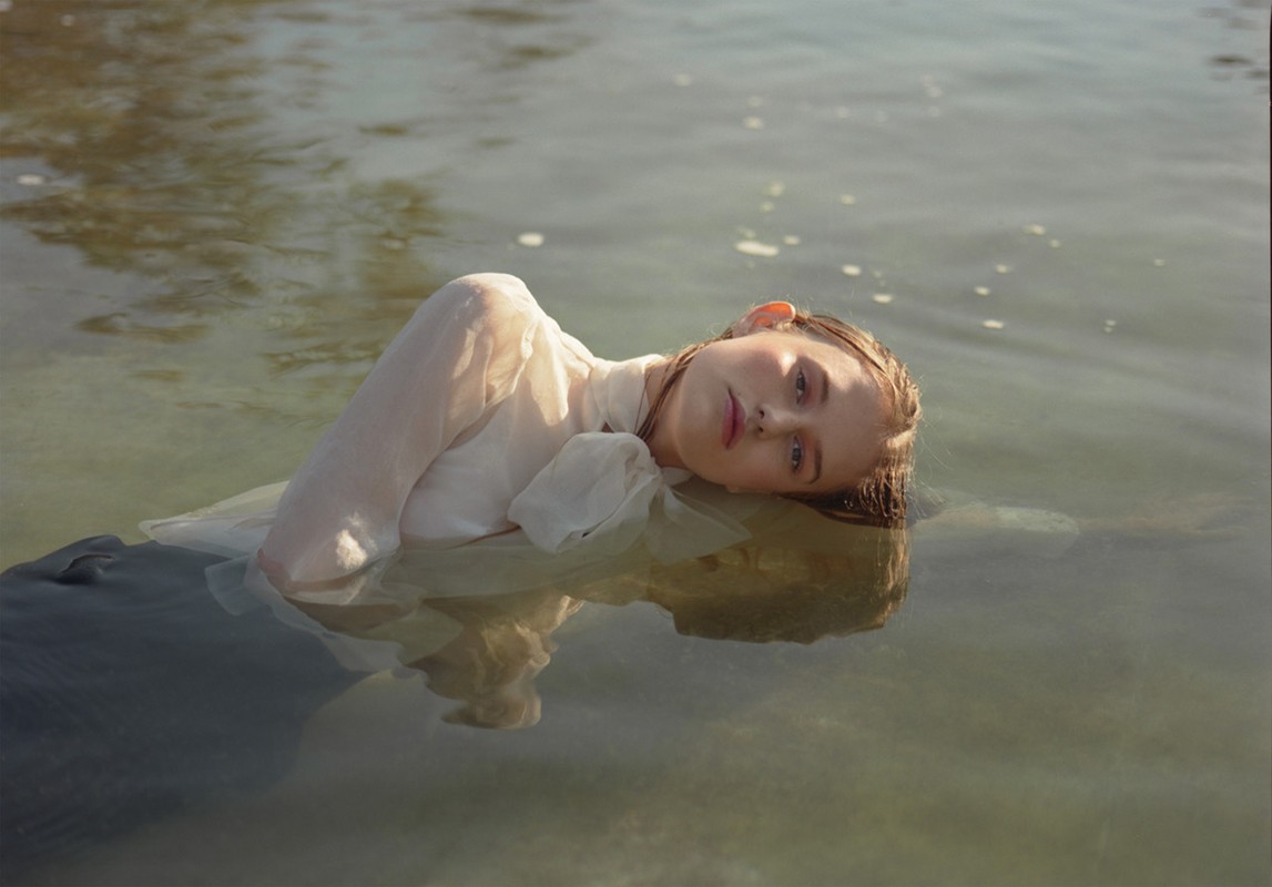 A woman in a white dress is lying in the water, her head above the surface, in an outdoor shot emphasizing tranquility and natural light.