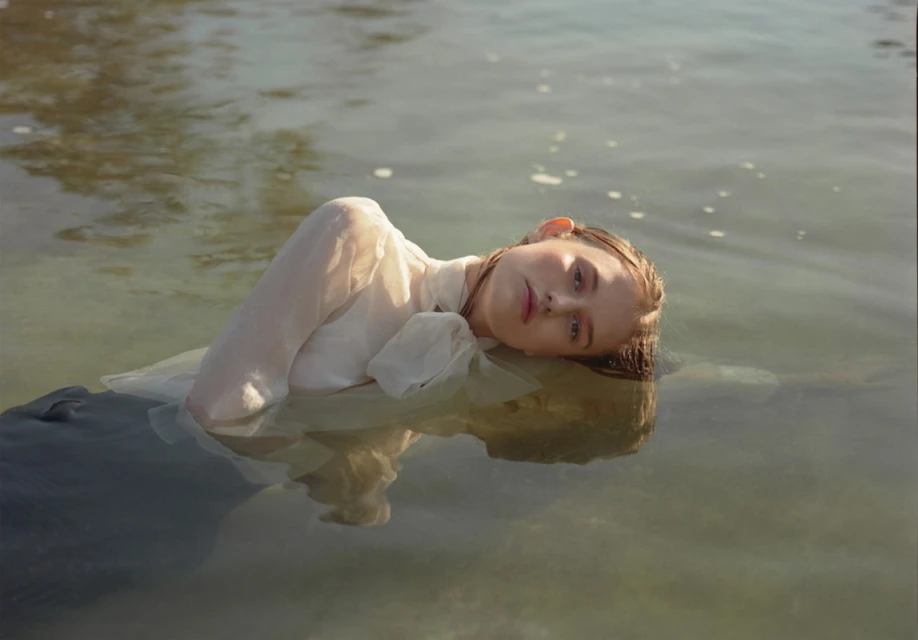 A woman in a white dress is lying in the water, her head above the surface, in an outdoor shot emphasizing tranquility and natural light.