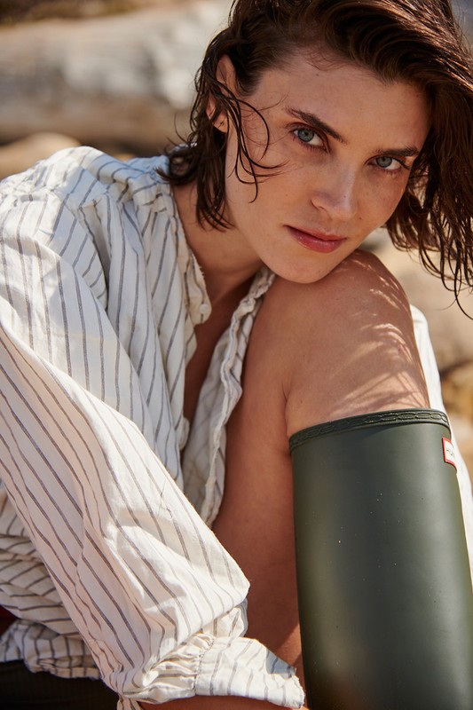 Adult woman with short hair wears a striped shirt and green rain boot posing outdoors in soft light on a rocky beach.