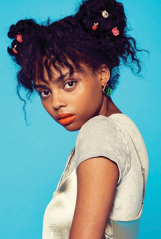 Portrait of a woman with an updo and orange lipstick, wearing a shimmering dress against a solid blue background, in a studio setting.