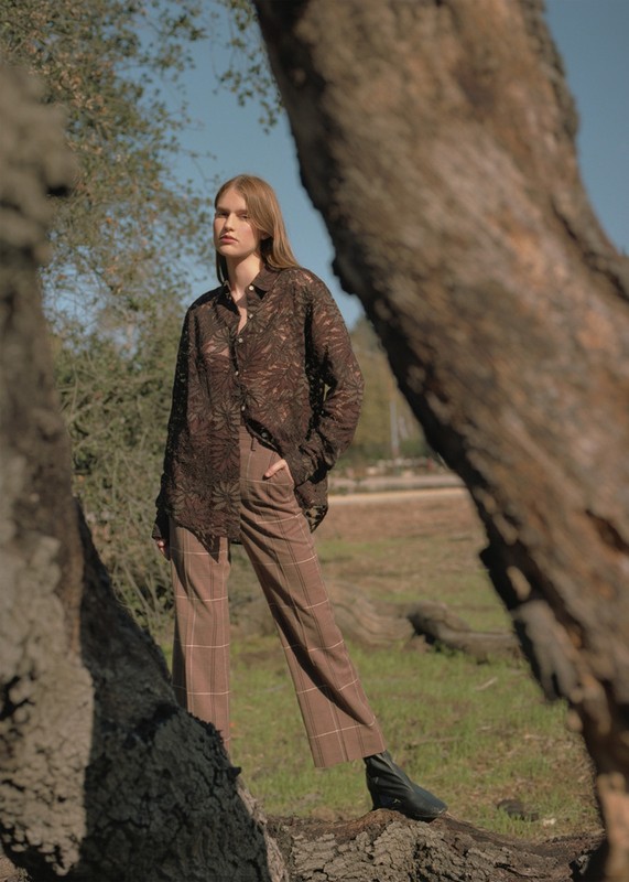 Woman in brown lace shirt and plaid pants stands in a field framed by tree trunks under a blue sky on a sunny day.