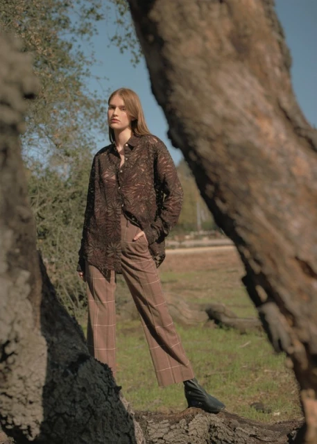 Woman in brown lace shirt and plaid pants stands in a field framed by tree trunks under a blue sky on a sunny day.