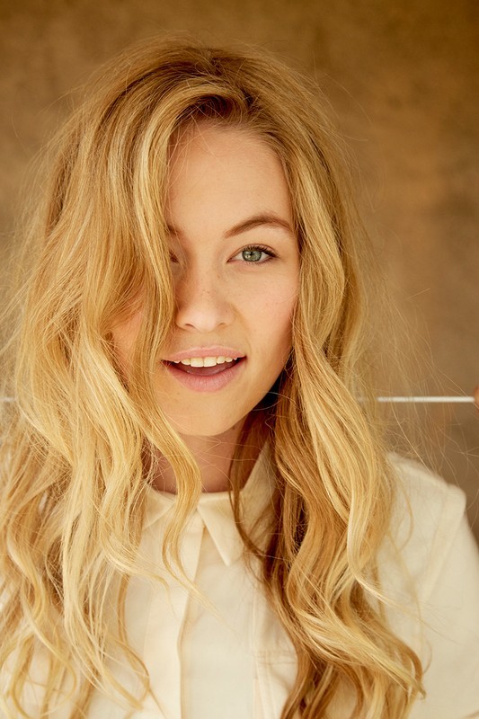Close-up portrait of a smiling woman with long blonde hair wearing a light-coloured shirt against a textured background.