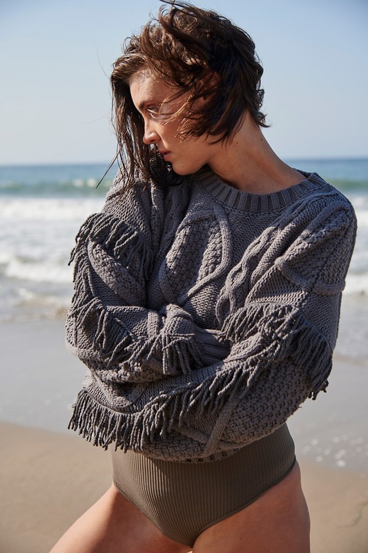 Woman in a grey knitted sweater with fringe and a swimsuit bottom poses outdoors on a beach with ocean waves in the background.