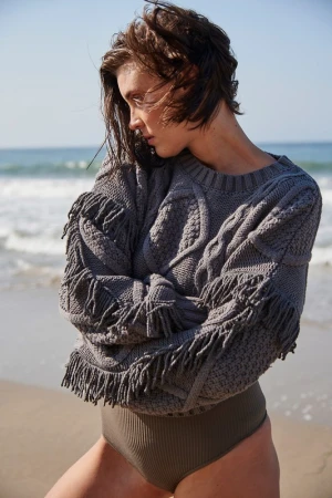 Woman in a grey knitted sweater with fringe and a swimsuit bottom poses outdoors on a beach with ocean waves in the background.