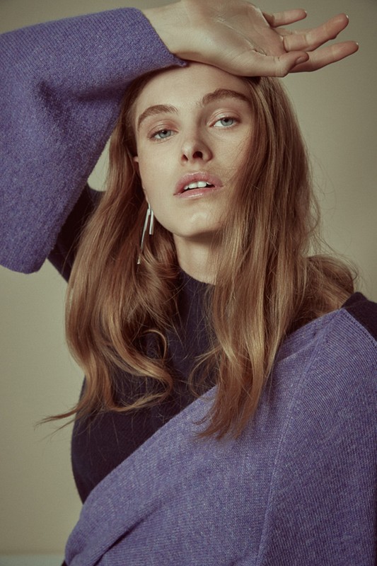 Portrait of a young adult woman with blonde hair, wearing a black top and purple sweater with long, wide sleeves against a neutral background.