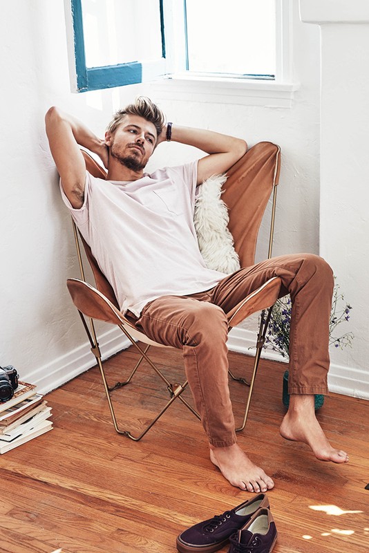 A blond man relaxes in a chair with a white t-shirt and brown pants in a well-lit indoor setting, with books and shoes on the floor.