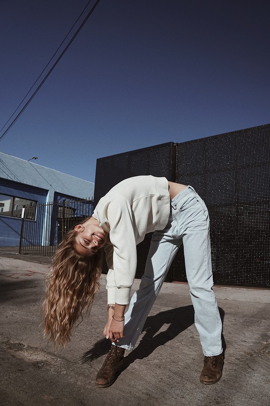 Young woman with blonde hair bends backward wearing jeans and a white sweater against an urban outdoor backdrop with a blue building.