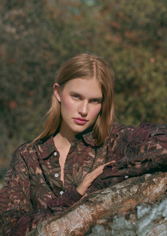 Blonde woman in patterned button-down poses outdoors with blurred forest background, resting her arm on a log.