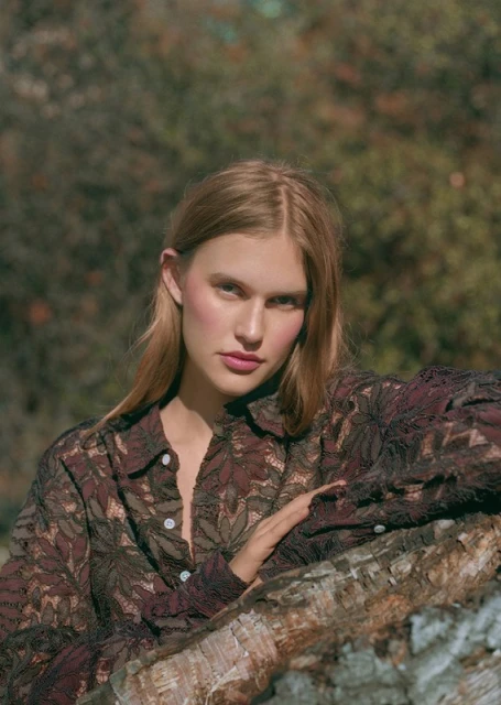 Blonde woman in patterned button-down poses outdoors with blurred forest background, resting her arm on a log.