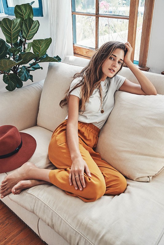 Woman with long hair and orange pants relaxing on a sofa near a window and potted plant with a hat, looking at the camera.