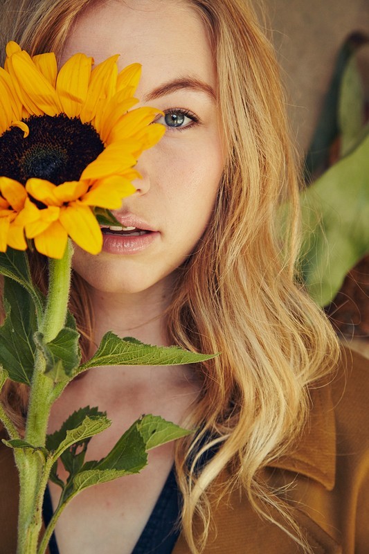 Close-up portrait of a woman with blonde wavy hair holding a bright yellow sunflower covering one eye in an indoor environment.
