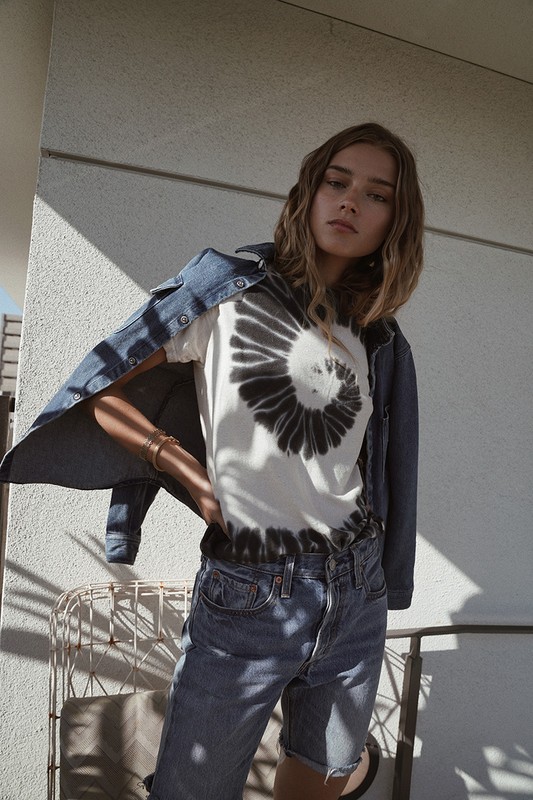 Blonde woman poses in a white t-shirt with a black spiral design, blue jean shorts, and a denim jacket against a neutral wall.