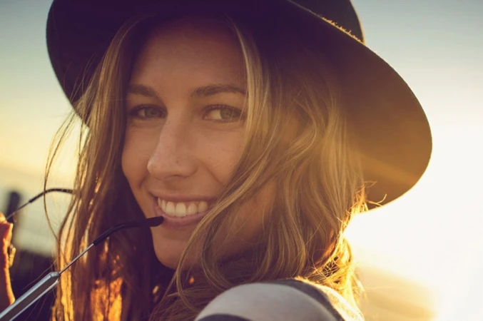 Close-up portrait of a smiling woman wearing a black wide-brim hat outdoors during sunset with warm backlight.