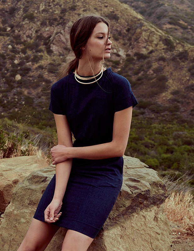Woman in dark navy blue dress wearing layered gold choker necklace seated outdoors on a mountain rock.