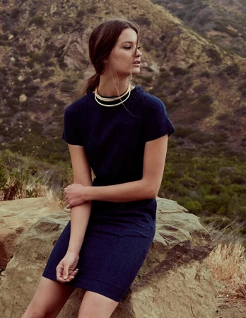 Woman in dark navy blue dress wearing layered gold choker necklace seated outdoors on a mountain rock.