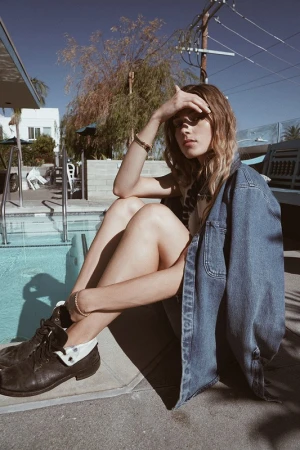 Woman sitting on ground next to swimming pool wearing a denim jacket and boots, shielding her eyes from the sun.