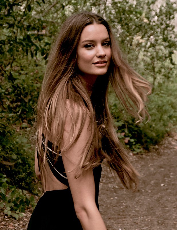 A young adult woman with long brown hair turns to look at the camera, standing on a path surrounded by green trees and foliage.