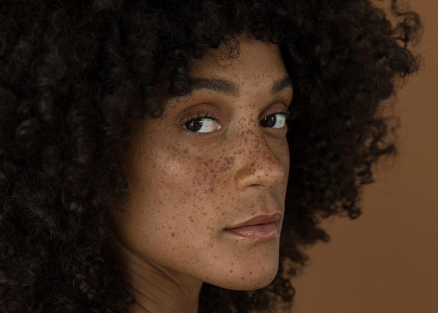 Close-up portrait of a woman with freckles and curly hair against a brown background, showcasing a profile view with soft natural lighting.