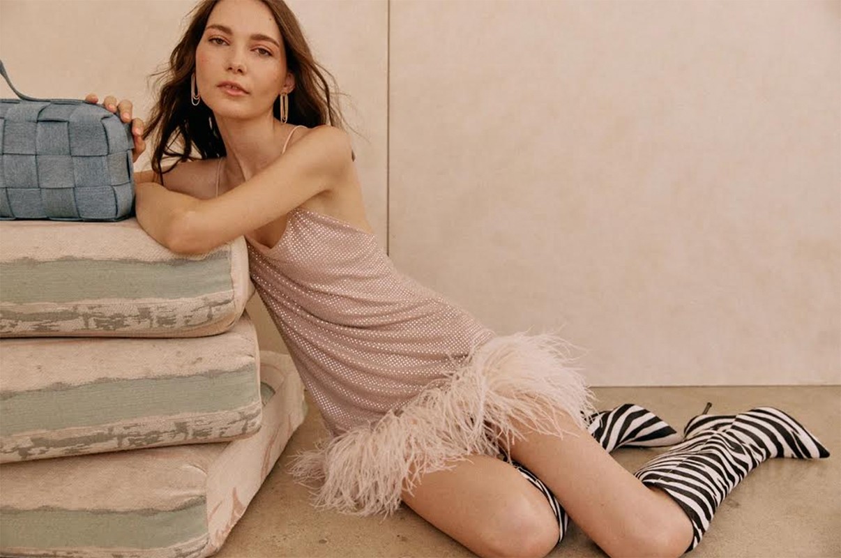A woman in a sparkling dress and zebra-striped boots poses next to a blue purse on stacked cushions in a fashion shot.