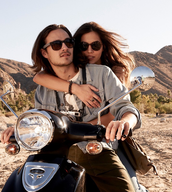 A couple rides a black motor scooter on a sunny day, set against a desert landscape with mountains, both wearing sunglasses.