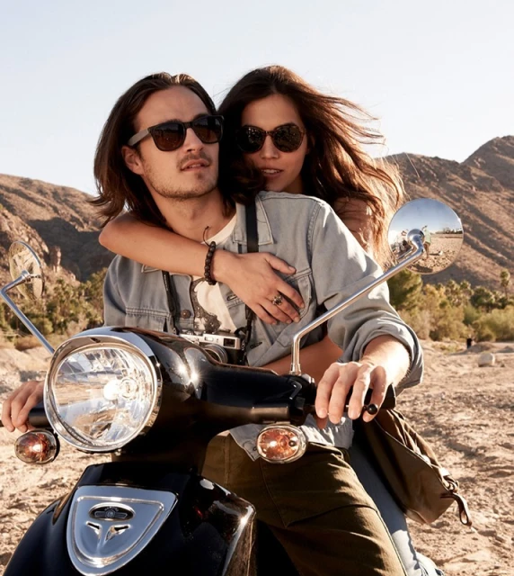 A couple rides a black motor scooter on a sunny day, set against a desert landscape with mountains, both wearing sunglasses.