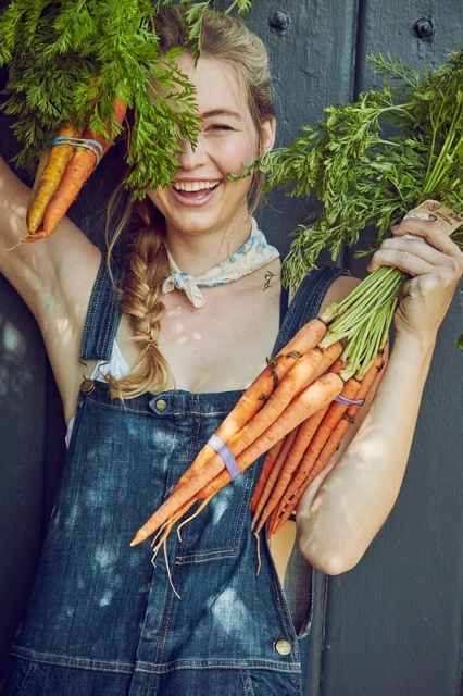 A smiling young woman in denim overalls holds up bunches of fresh carrots against a dark gray wooden background in soft lighting.