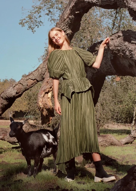 A fair-skinned woman in a pleated green dress poses with a goat under a large tree on a sunny day in an outdoor, grassy environment.