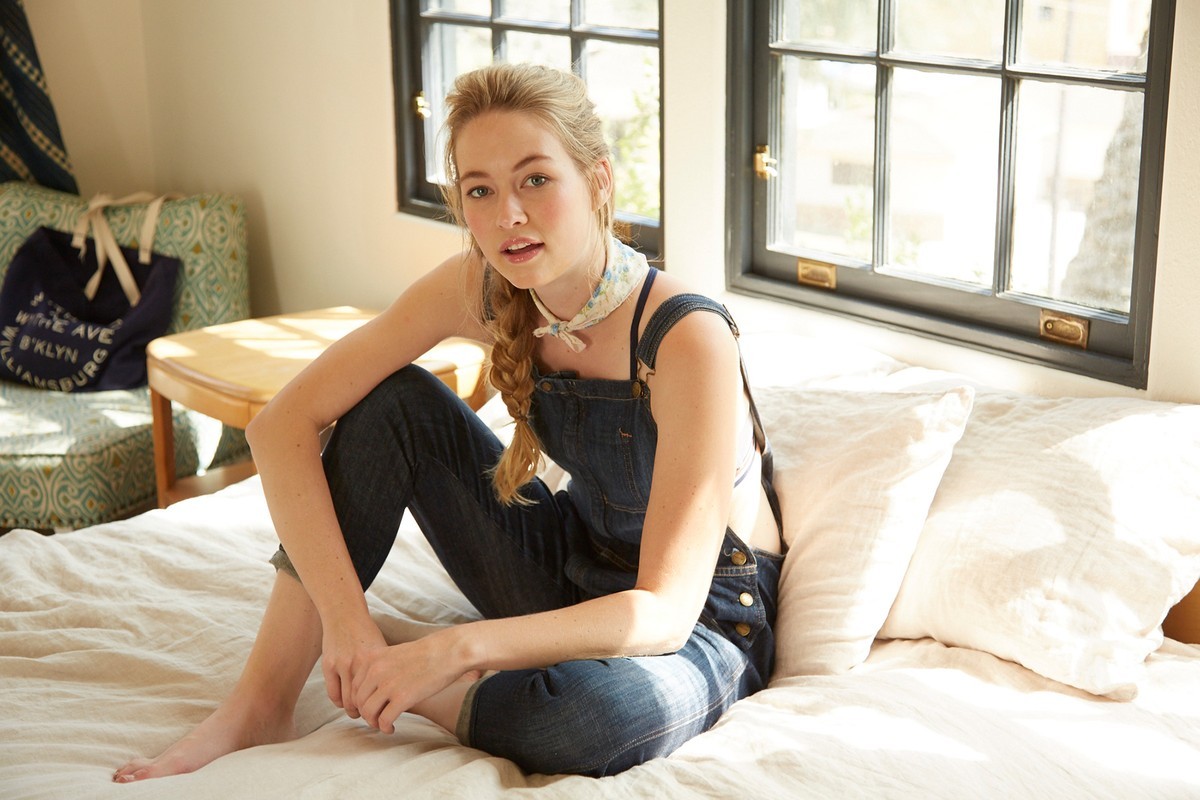 A young adult female poses indoors on a bed with natural lighting, wearing denim overalls and a patterned neck scarf, looking at the camera.
