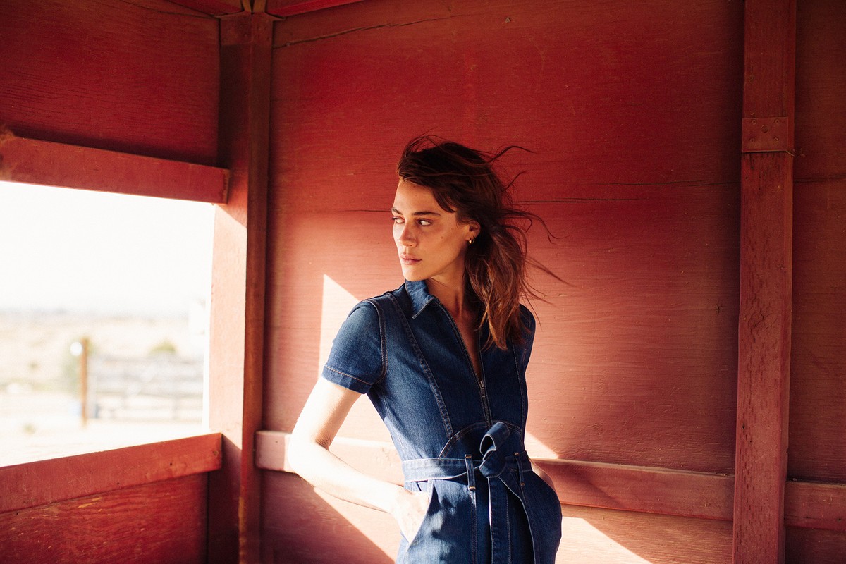 An attractive adult woman stands inside of a red-painted wooden structure, wearing a denim jumpsuit with a sash, gazing out into the landscape.
