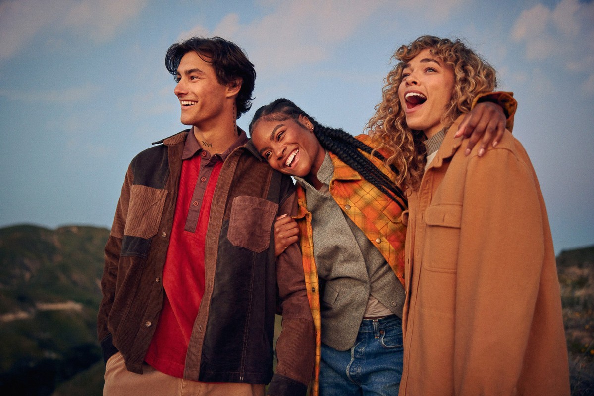 Three smiling friends wearing vintage outerwear pose outdoors with a mountain backdrop.  The group is casually dressed.