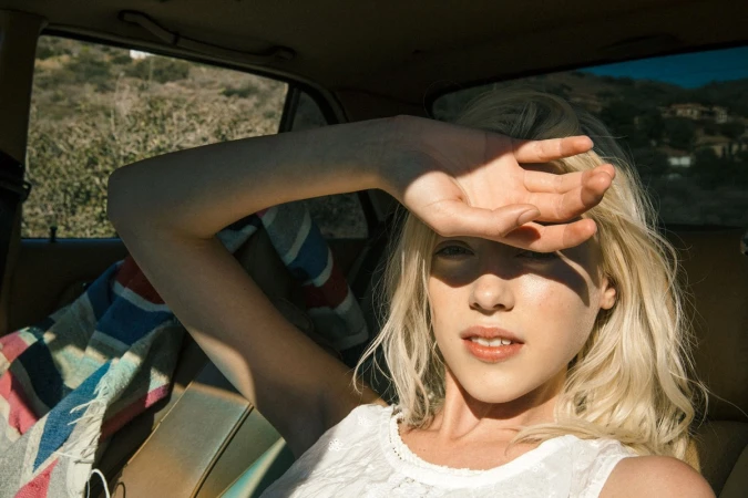 Blonde woman sitting inside a car with her arm raised shielding her eyes from bright sunlight in Los Angeles.
