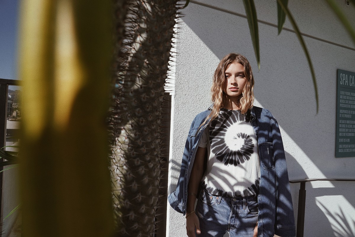 A young fair-skinned woman stands outside, wearing a tie-dye shirt and denim jacket, partially obscured by greenery on a sunny day.